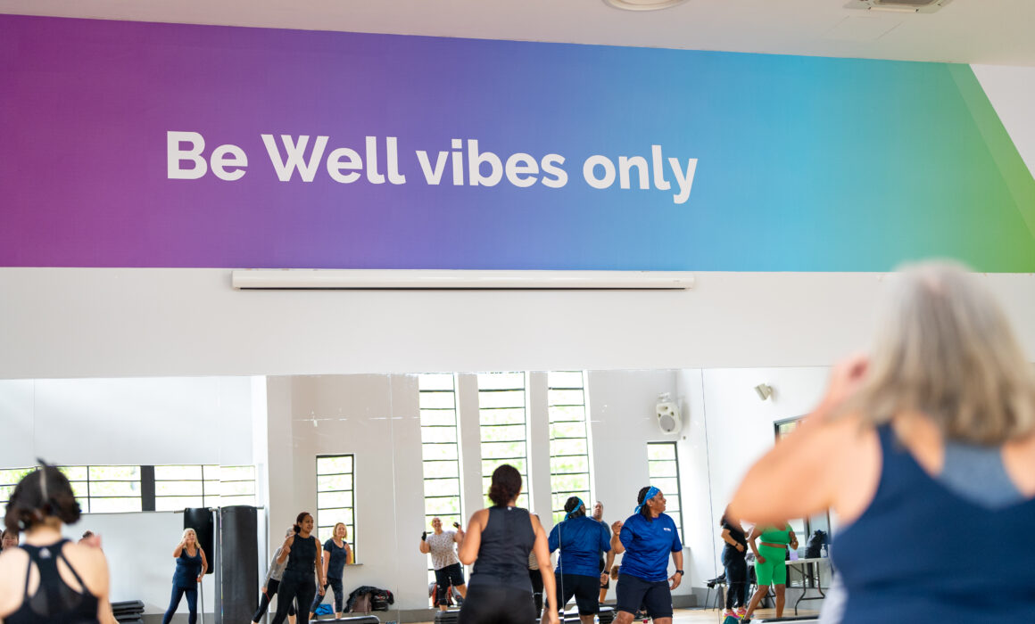 A photo of some people participating in a fitness class. There's a colourful sign in the bavkground that says 'Be Well The Spa'