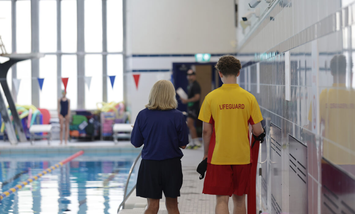 A photo of a lifeguard standing my a pool with his back to the camera. His tshirt is yellow and says lifeguard. He stands next to a Be Well staff member who is also next to the swimming pool