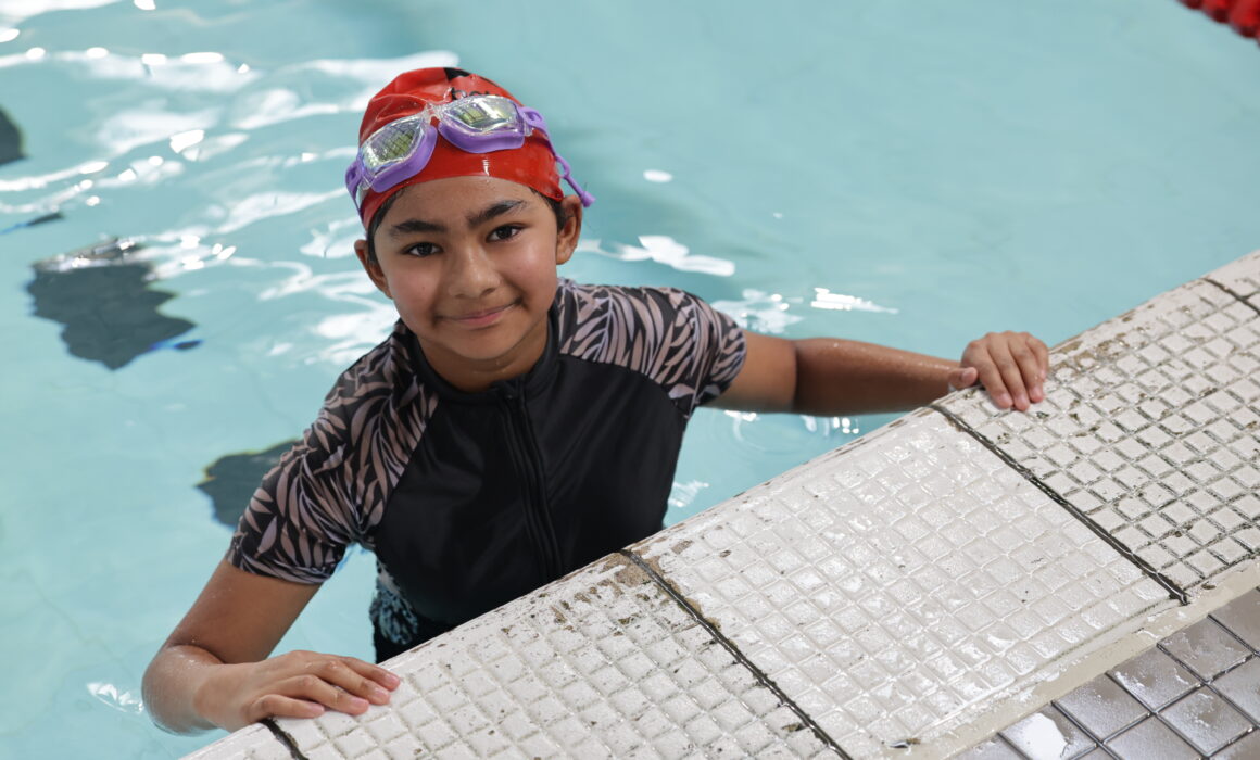 A photo of a young asian girl at the edge of a swimming pool. She wears a googles, swim hat and swim suit.