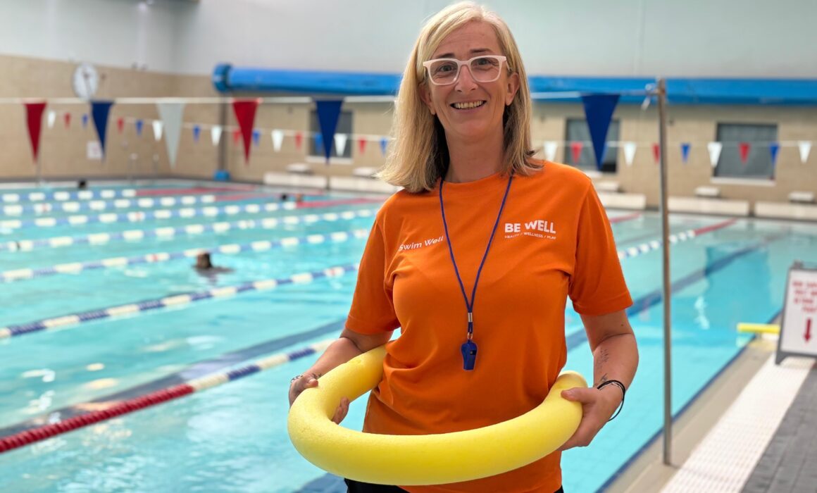 A swim buddy wears and orange t-shirt whilst holding a pool noodle in front of a swimming pool