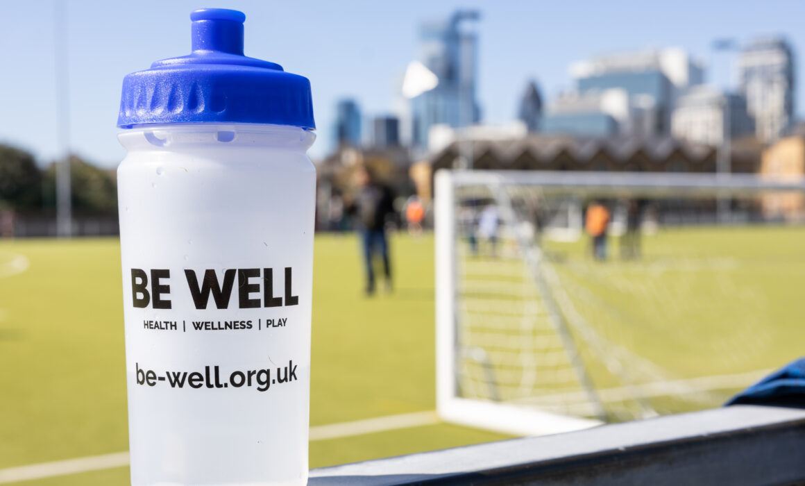A photo of a translucent Be Well branded water bottle by a football pitch