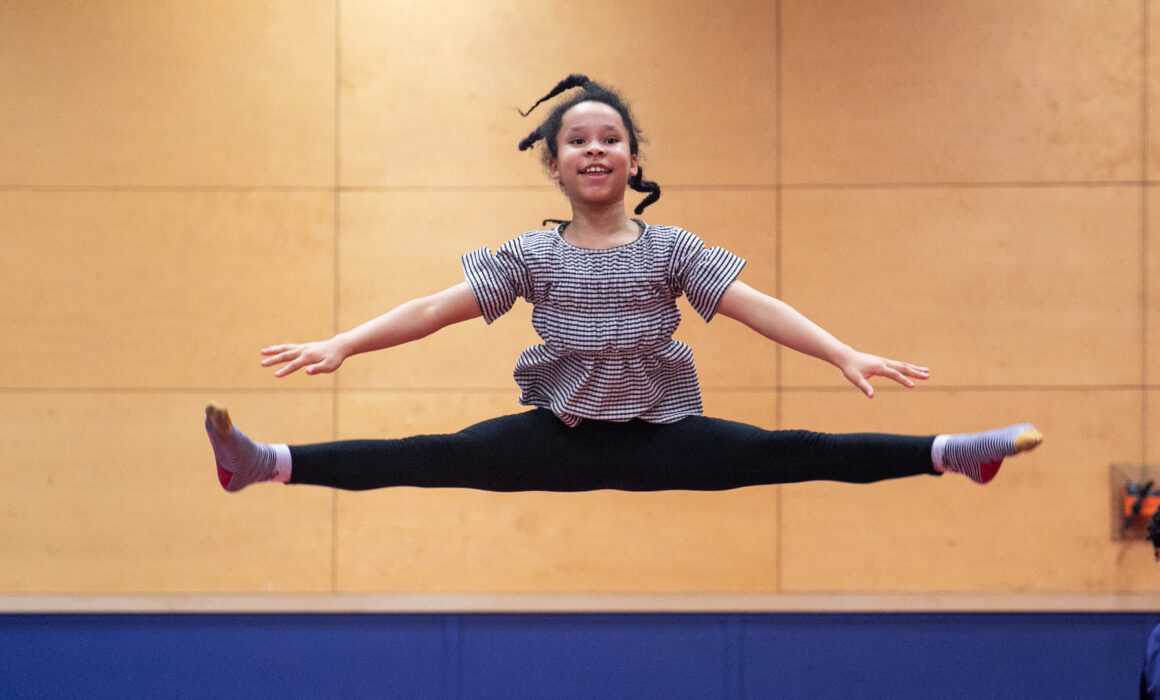 A photo of a young girl performing a gymnastics straddle jump. She smiles as she performs the jump