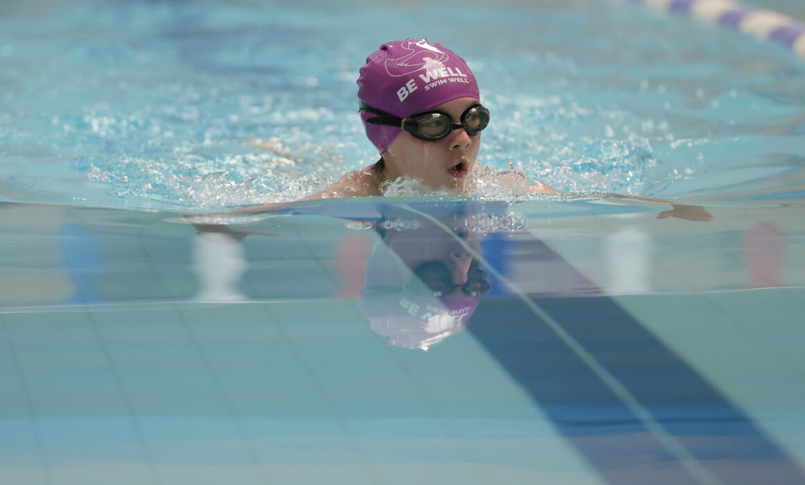 A young girl swims in a swimming pool. She is wearing a purple swimming hat that says 'Be Well Swim Well'