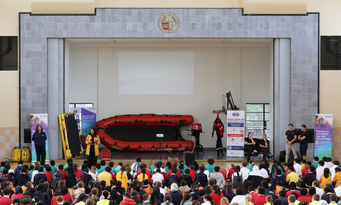 A photo of many school children sat in front of a stage at Poplar Leisure Centre for a swim safety event. On stage is an inflated lifeboat as well as speakers from Swim England, The London Fire Brigade, The Marine Police and Be Well Swimming staff.